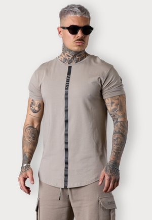 Beige T-Shirt mit kurzen Ärmeln und einem schwarzen vertikalen Streifen, versehen mit Logo-Text. Das Modell trägt eine Sonnenbrille und zeigt komplexe Armtattoos.