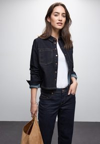 Giacca cropped in denim scuro con due tasche frontali, chiusura con bottoni e polsini azzurri. Abbinata a jeans scuri e una borsa in suede beige.