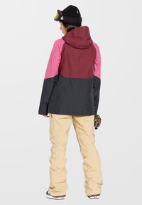 Geïsoleerde jas in zwart, bordeauxrood en roze, met capuchon en een relaxte pasvorm. Gecombineerd met beige broek, met cargozakken en laarzen.