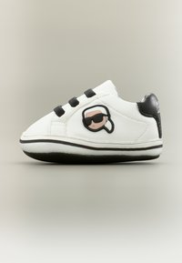 Λευκό παιδικό αθλητικό παπούτσι με μαύρα κορδόνια, μαύρη ταμπέλα στη φτέρνα και λογότυπο καρτούν με πρόσωπο που φοράει γυαλιά ηλίου στο πλάι.