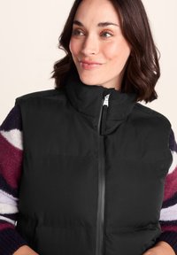 Woman with short brown hair wearing a black zip-up puffer vest over a maroon, purple, and white striped sweater, looking slightly right and smiling.
