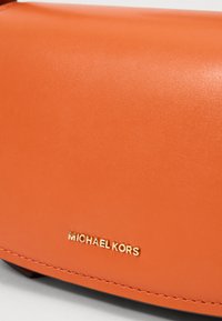 Oranžna usnjena torbica z gladko teksturo, ki vsebuje zlato blagovno znamko "MICHAEL KORS" in poenostavljen dizajn. Vidni šivi.