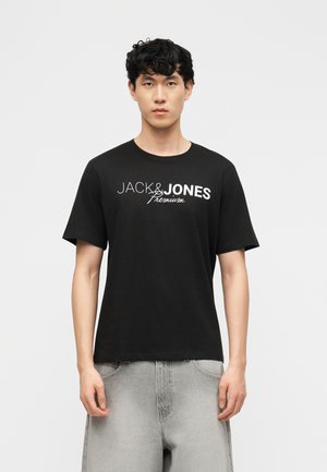 Noor mees lokkis mustade juustega, seljas must "JACK & JONES Premium" T-särk ja heledat halli värvi teksad, seisab ühevärvilise tausta ees.