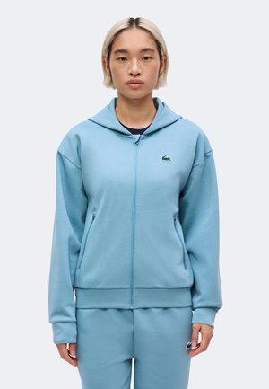 Persona joven con cabello corto y claro lleva una sudadera con capucha azul claro con cremallera y pantalones deportivos a juego con el logo de cocodrilo en el pecho y la cadera.