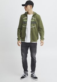 Sudadera verde con cremallera y texto en la parte delantera, camiseta blanca debajo, jeans oscuros deslavados y zapatillas negras con detalles en blanco.