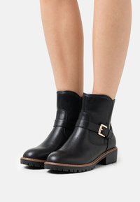 Las botas de tobillo de cuero negro presentan una puntera redondeada y una correa con un hebilla en tono dorado. La suela texturizada ofrece agarre y estabilidad.