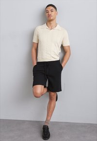 Cremefarvet strikket polo med struktureret mønster, parret med sorte shorts og sorte sneakers. Modellen poser med det ene ben korslagt og hænderne i lommerne.