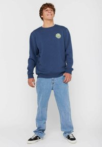 Navy blue sweatshirt with a round, green graphic logo on the chest, paired with light blue denim jeans and black and white sneakers.