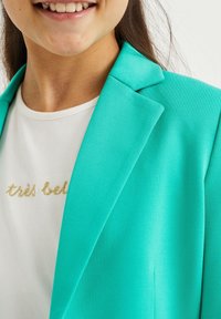 Tyrkysový oblekový blejzr s hladkou texturou, s klopovým límcem a přiléhavým střihem, přehozený přes bílou košili se zlatým textem.