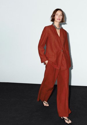 Roestkleurig linnen pak met een blazer met één knoop en brede pijpen. Model draagt flip-flops en een decoratieve ketting.