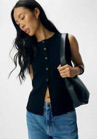 Schwarzes ärmelloses Top mit Knöpfen, das ein gespaltenes Design aufweist. Kombiniert mit blauen Jeanshosen und einer schwarzen Leder-Schultertasche.