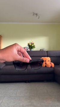 Óculos de sol pretos com lentes escuras em degradê, armações quadradas e discretos detalhes em metal, segurados à mão. Mobiliário suave ao fundo.