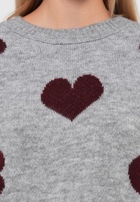 Sivi pleteni pulover s burgundskim uzorkom srca u središtu, teksturirana tkanina s okruglim izrezom i rebrastim detaljima.