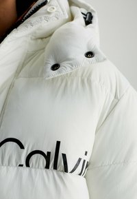 Bílá péřovka s matným povrchem, se znakem velkého černého "Calvin", kulatými černými patentkami a designem s vysokým límcem. Měkká textura.