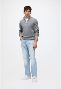 Jeune homme portant un pull gris à fermeture éclair quart de zip, jean bleu clair, baskets blanches, debout avec une main dans la poche devant un fond blanc uni.