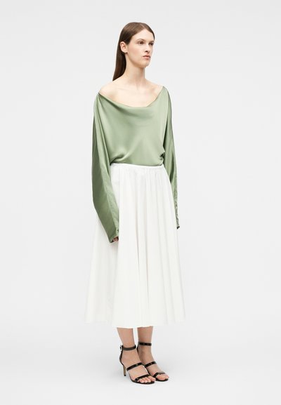 Γυναίκα όρθια, φορώντας μια χαλαρή σατέν μπλούζα σε πράσινο φασκόμηλο με ανοιχτούς ώμους, λευκή μεσαίου μήκους φούστα και μαύρα πέδιλα με ψηλό τακούνι και λεπτά λουράκια.