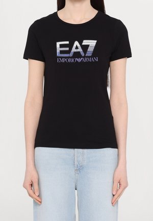 Vrouw draagt een zwart T-shirt met korte mouwen met het "EA7 Emporio Armani"-logo in blauw en wit, gecombineerd met lichtblauwe jeans.