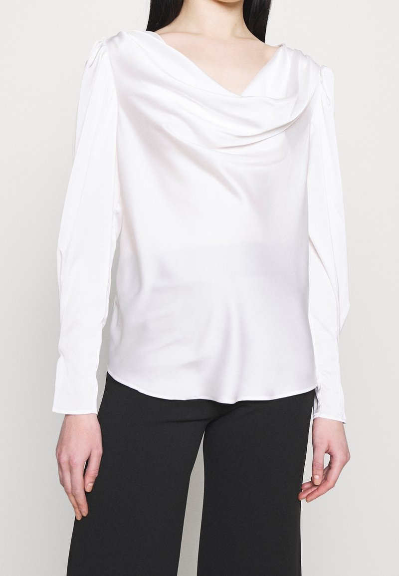 Blouse en satin blanc avec un col bénitier et des manches longues, présentant une texture lisse et une coupe décontractée, portée sur un pantalon noir.