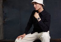 Chemise noire à manches longues avec boutons, chapeau seau blanc, pantalon de couleur claire, avec une montre argentée à cadran noir et bracelet transparent.