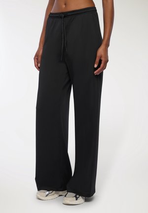 Femme portant un pantalon noir ample avec taille à cordon et des chaussures de sport beiges, debout devant un fond clair uni.