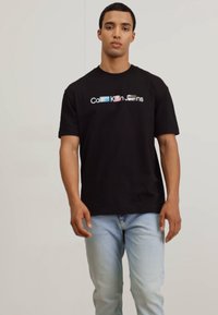 Camiseta de algodón negra con un logo blanco y acentos coloridos. Combinada con jeans azules claros, utilizada por un modelo masculino de pie.