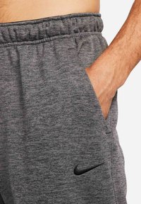 Main dans la poche d'un pantalon de survêtement gris Nike avec taille élastique et logo Swoosh noir sur la cuisse.