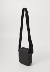 Borsa a tracolla nera di forma quadrata, realizzata in materiale morbido. Presenta una chiusura con zip e una tracolla in tessuto con il nome del marchio in rilievo.