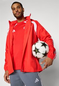 Muž v červenom športovom saku a košeli drží pod paľou bielo-čiernu futbalovú loptu so vzorom hviezd na jednoduchom pozadí.