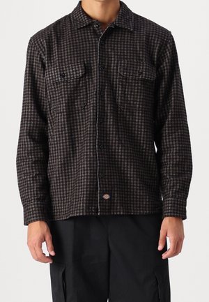 Homme portant une chemise à carreaux marron foncé et noire boutonnée, avec deux poches poitrine, et un pantalon noir.
