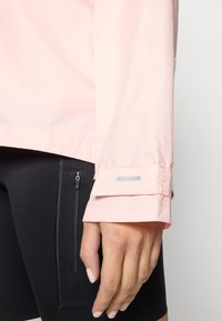 Light pink waterproof jacket with a textured finish, featuring adjustable cuffs and a reflective accent, paired with black form-fitting shorts.