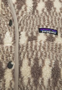 Tela de forro polar con un patrón marrón y crema, con una superficie texturizada, que presenta un logo negro de Patagonia y un ribete gris a lo largo de los bordes.