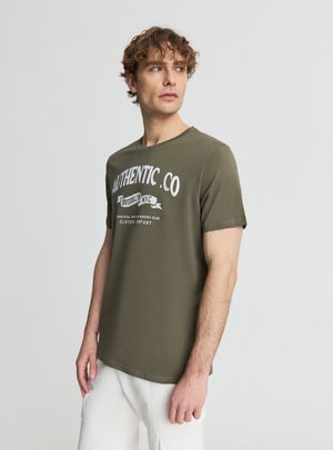 Młody mężczyzna z kręconymi włosami, ubrany w oliwkowozieloną koszulkę "Authentic Co" i białe spodnie, spoglądający lekko na bok, na jednolitym tle.