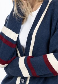 Marinblå cardigan med röda och krämfärgade ränder, som har en ribbad krage och knappdetaljer. Mjuk textur med avslappnad passform och öppen framsida.