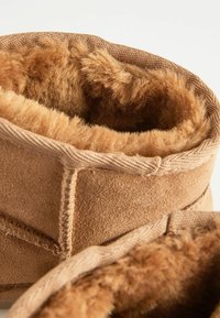 Bruna mocka stövelkanter med en mjuk, fluffig beige fleecefoder. Har stickade sömmar och en strukturerad yta för hållbarhet.