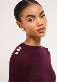 Burgundská rebrová sveter s okrúhlymi striebornými gombíkmi na pleci, s vysokým výstrihom a štrukturovanou tkaninou.