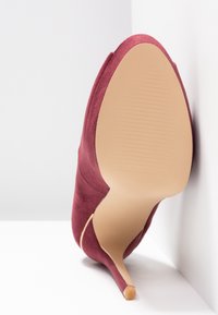 Chaussure à talon haut en daim bourgogne avec un bout pointu et une silhouette épurée. La semelle est beige lisse avec des stries subtiles pour une meilleure adhérence.