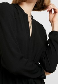 Schwarzes Langarmkleid mit V-Ausschnitt und lockerer Passform. Verfügt über einen weichen, strukturierten Stoff und gestapelte silberne Halsketten.