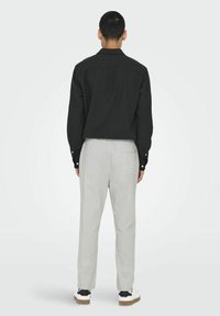 Camisa negra de botones con cuello, combinada con pantalones grises claros que presentan una cinturilla elástica y un bolsillo trasero. Los zapatos tienen una base blanca con detalles en negro.