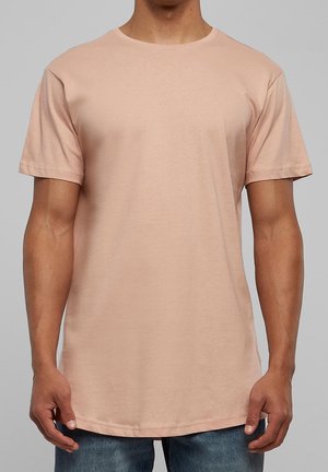 Mann trägt ein schlichtes, kurzärmeliges hellbeiges T-Shirt und blaue Jeans, steht vor einem neutralen grauen Hintergrund.