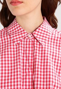 Chemise boutonnée en vichy rouge et blanc avec un col classique, présentant une texture en coton lisse et une patte de boutonnage centrale.