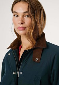 Marineblauwe jas met een bruine korduroy kraag, zilveren hardware, ritssluiting en zak. Soepele textuur met een casual, gestructureerd ontwerp.