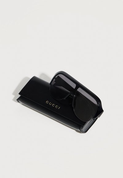 Schwarze übergroße Gucci-Sonnenbrille, die auf einem passenden schwarzen Etui mit goldenem Gucci-Logo vor hellem Hintergrund liegt.