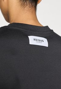 Černá bavlněná mikina s kulatým výstřihem a viditelnou bílou etiketu Adidas Sportswear na zadní straně. Hladká textura, ležérní design.