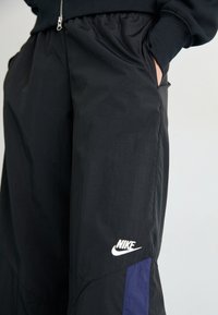 Svarta träningsbyxor med lös passform, elastisk midjeresor, fickor och en vit Nike-logotyp. Har en lila detalj på sidan.