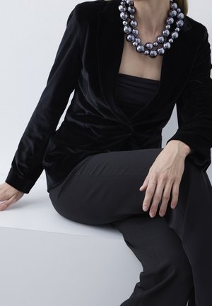 Frau in schwarzem Samtblazer und Hose sitzt, trägt eine klobige silberne Perlenkette, mit einer Hand auf dem Knie ruhend und der anderen auf der Oberfläche.