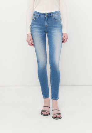 Femme portant un jean skinny bleu clair et des sandales noires à brides, debout devant un fond blanc.