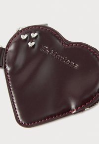 Δερμάτινη τσάντα σε σχήμα καρδιάς σε μαβί απόχρωση με ροζ ραφές, ασημένιες καρδιές και ανάγλυφο λογότυπο "Dr. Martens". Κλείνει με φερμουάρ στο πάνω μέρος.