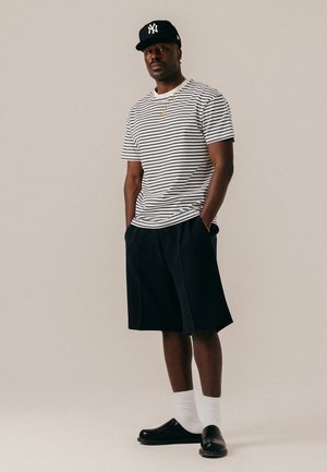 Homme debout, les mains dans les poches, portant une casquette noire des New York Yankees, un t-shirt rayé blanc et noir, un short noir, des chaussettes blanches et des chaussures noires.