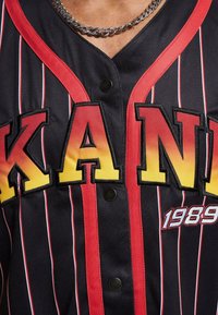 Svart trøye med røde detaljer, vertikale striper, som har fet, gradient gule bokstaver "KANYE" og året "1989." Svart knapper og V-hals design.