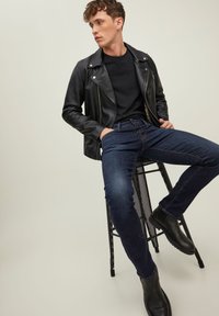 Mladic sjedi na crnoj metalnoj stolici, odjeven u crnu kožnu jaknu, crnu majicu, tamnoplave traperice i crne čizme, na bež pozadini.